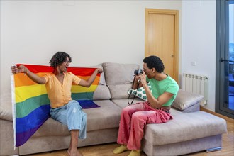 Gay man holding LGBT rainbow flag while boyfriend taking photos sitting on the sofa at home