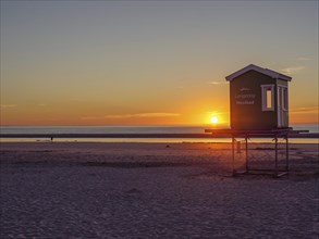 Beach at sunset with a raised lifeguard tower and a wide view of the sea and sky, setting sun on