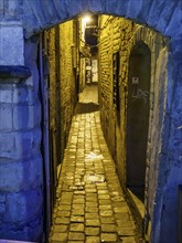 Narrow, cobbled alley between historic buildings, illuminated in warm light at night, blue hour in