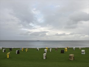 Wide green meadow with scattered colourful beach chairs, behind it the sea and cloudy sky,