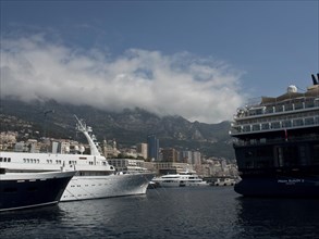 Yachts lying in the water against a backdrop of mountains and city buildings under a partly cloudy