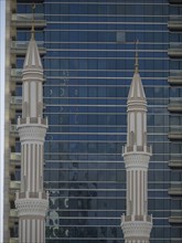 Two minarets of a mosque in front of a modern building with glass facade and sky in the background,