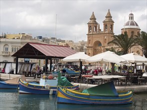 Boats and restaurants at the harbour with a church in the background on a cloudy day, Valetta,