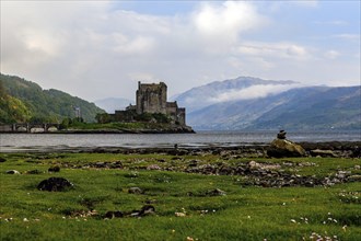 A castle on the lakeshore with mountains and clouds in the background, surrounded by green meadows