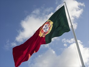 Portuguese flag in the colours green, red and yellow waving in the wind in front of a cloudy sky,