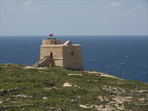 Historic watchtower on a rocky coast under a blue sky with a flag on top, green field with flowers