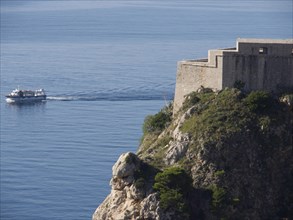 A historic fortress on a rocky cliff above calm blue waters, a boat passes by in the distance, the