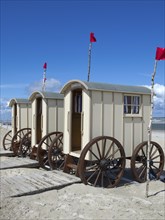 Three beige changing cabins with large wooden wheels on a beach under a blue sky with clouds,