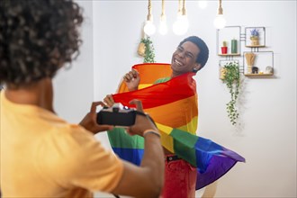 Gay man wrapping with lgtb flag while couple taking photos of him at home