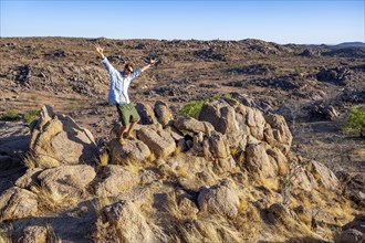 Young man stretching his arms in the air, barren landscape with rocky hills and acacia trees,