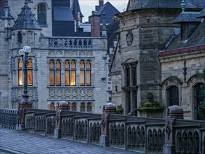 Gothic buildings with illuminated windows and an old stone bridge at dusk in a historic town,