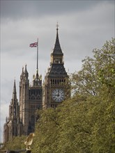A tall clock tower with a British flag on a cloudy sky and surrounded by green trees, London,