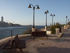 Seafront promenade with benches and decorative lamps, paved floor and railings under a clear sky,