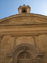 Facade of a historic church with an ornate bell tower under a clear blue sky, the town of mdina on