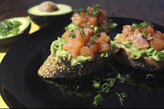 Three slices of seeded bread topped with avocado, salmon, and dill on a black plate with avocado