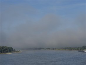 Blue sky over a calm, misty river with wooded shorelines, the Rhine at Wesel with a ruined bridge