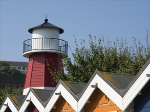 Colourful lighthouse and houses with pointed roofs in front of a clear sky and green trees,