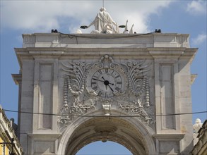 Ornate triumphal arch with central clock and sculptures, against a blue sky, Lisbon, Portugal,