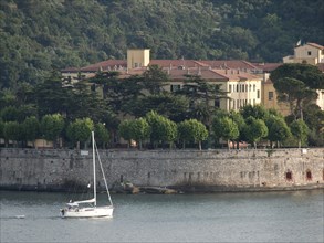 Yellow building on the water with a sailing boat in the foreground, surrounded by green trees and