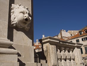Close-up of a stone sculpture of a lion's head in front of historic buildings under a clear blue