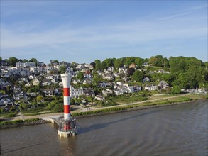 Red and white lighthouse on the riverbank surrounded by houses and trees under a clear sky, houses