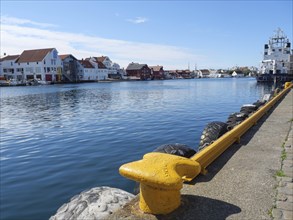 Waterfront with a yellow bollard and tyre protection, water and city in the background, in sunny