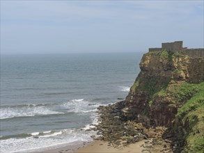 A rocky coastline with waves crashing on the beach and a castle ruin on the cliffs, old ruin by the