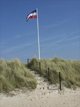 A staircase leads through grassy dunes to a high-flying flag, the sky is clear and blue,