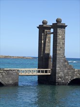 Two old stone towers connect with a small bridge over the water under a clear sky, the Canary