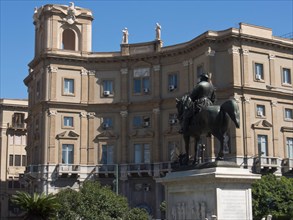 Equestrian statue of a historical figure in front of a classical building with statues, on a clear