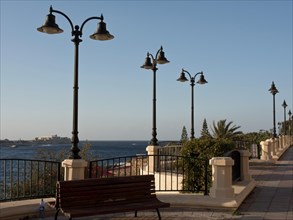 Promenade along the sea with benches, decorative lamps and palm trees, railings and clear sky,