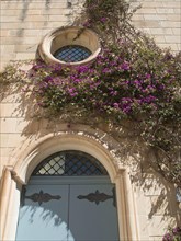 A blue door with an arch and a round window surrounded by ivy and purple flowers on a stone wall,