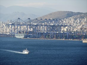 A large harbour with ships and containers, behind it a city and mountains, ancient buildings with