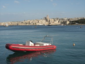A red inflatable boat floats calmly on the sea in front of the city with historic buildings in the