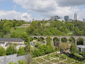 Urban landscape with a viaduct, modern buildings and wooded hills under a cloudy sky, overlooking a
