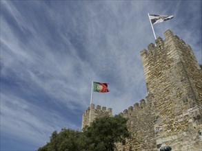Two towers of a castle with waving flags and surrounded by trees under a blue sky, Lisbon,