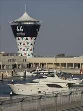 A tall tower with a striking pattern stands in the harbour with yachts and other boats, buildings