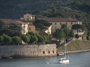 Yellow building on the waterfront, a sailboat in the foreground and forest covered hills in the