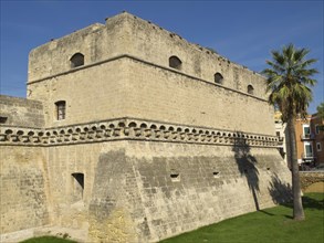 Old fortress with imposing stone walls and a palm tree in the foreground under a bright blue sky,