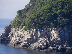 Rocky coast with densely overgrown trees and a small lighthouse overlooking the calm blue sea, the