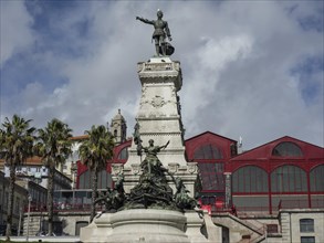 Monument with statue in front of a building with a red roof under a cloudy sky, historic buildings