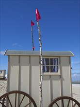 Side view of a beige changing cubicle with wooden wheels and two red flags under a blue sky,