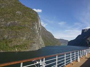 View from a ship's deck of steep mountains and a wide fjord, watched over by clear blue skies,