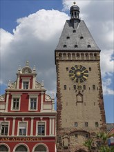 Historic tower with large clock and a red building in the foreground under blue sky with clouds,