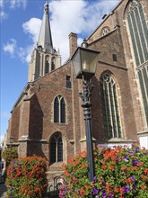 Historic red brick gothic church with high steeple, a large flower bed and lamppost under a blue
