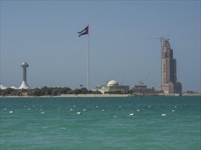 A flag flies over coastal buildings with a domed building and towers, the sea glistens intensely