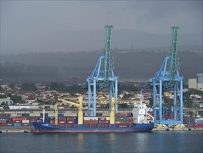 A freighter stands in the harbour with large blue cranes and many containers in the background,