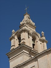 A large stone church tower with architectural details rising into the blue sky, the town of mdina