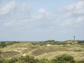 Hilly dune landscape with a view of distant trees and the sea under a cloudy sky, Spiekeroog,