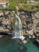 A waterfall cascades from a cliff into turquoise-coloured water in a rocky, natural setting, aerial